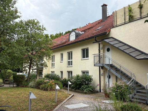Helles Gebäude mit roten Dach, Garten und Treppe im Freien, umgeben von Bäumen.